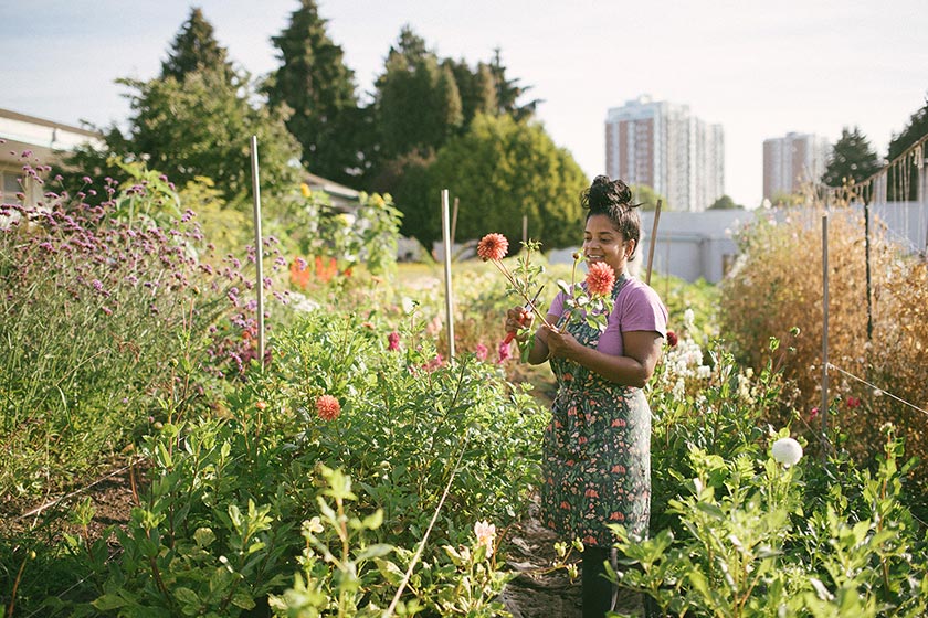 A woman tending to a community garden with a city in the background.